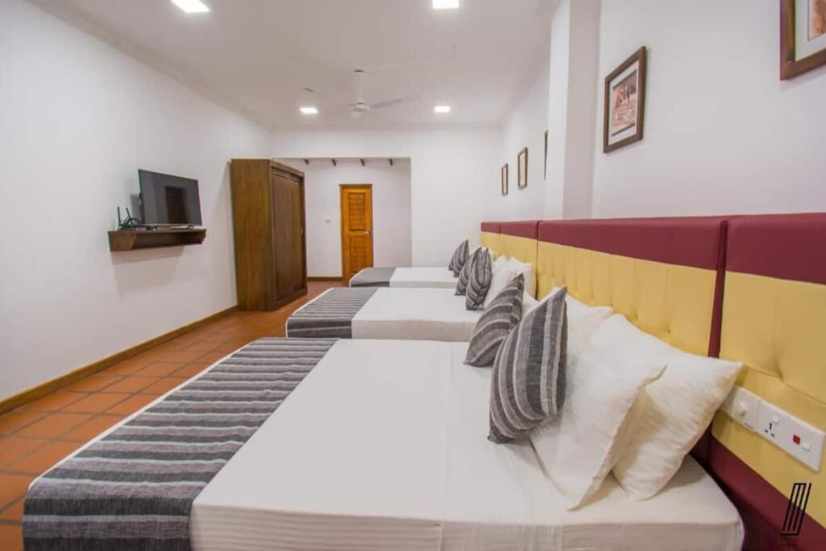 Une chambre d’hôtel avec quatre lits alignés en rangée, un téléviseur posé sur une étagère, une armoire et une porte visible au fond.