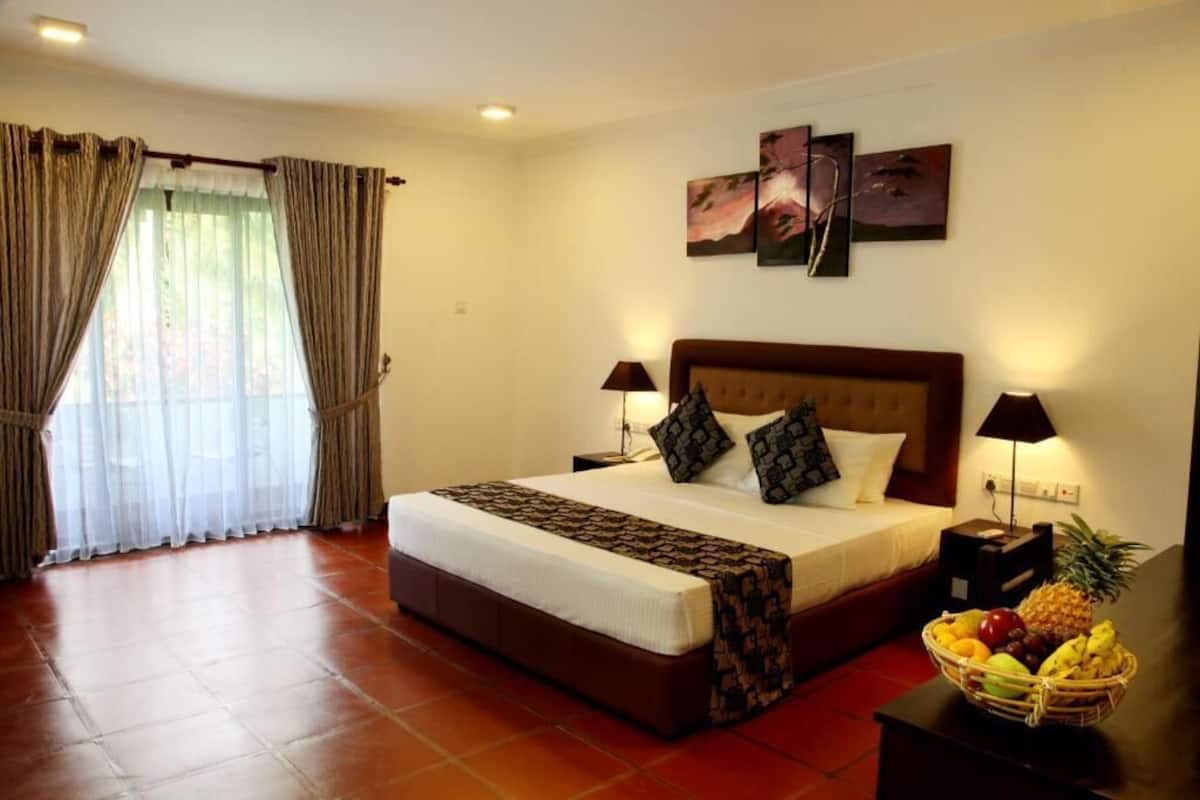 Une chambre d’hôtel avec un grand lit, deux lampes de chevet, un panier de fruits et une vue sur l’extérieur à travers des rideaux légers.