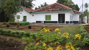 A white building with a red-tiled roof and green shutters, surrounded by a garden with various plants and flowers. - Green Banana Mirissa (Mirissa)