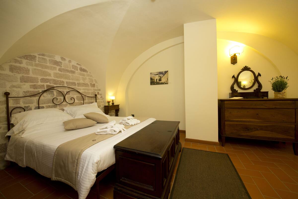 A bedroom with a stone wall, a bed with white linens, a wooden dresser, and a mirror.