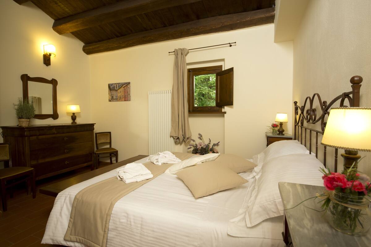 A hotel room with a bed, bedside tables, a dresser, a mirror, and a window with a view of greenery.