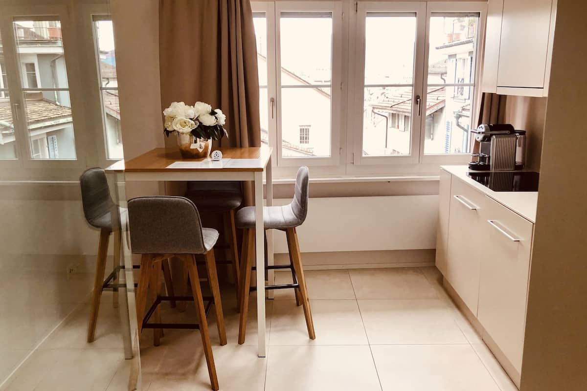Una pequeña cocina con una mesa de comedor, dos sillas, una cafetera y un ventanal con cortinas.