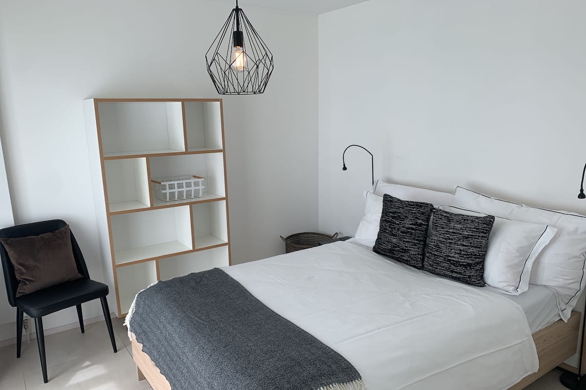 Un dormitorio con cama, una silla, estantería e iluminación colgante.