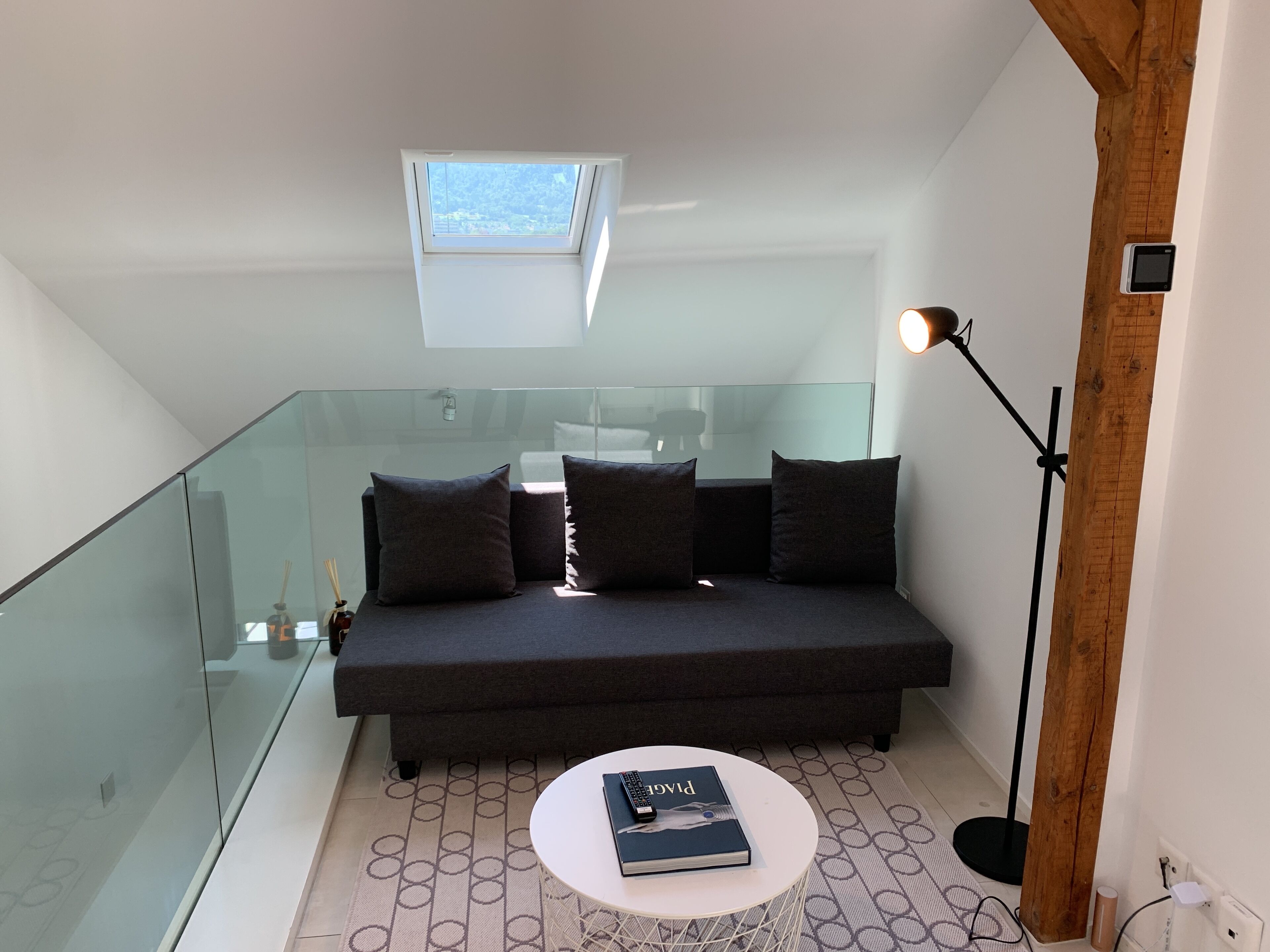 Ein modernes Schlafzimmer im Loft-Stil mit einem Glasbalkon, einem dunklen Sofa, einem kleinen runden Tisch und einer Deckenleuchte.
