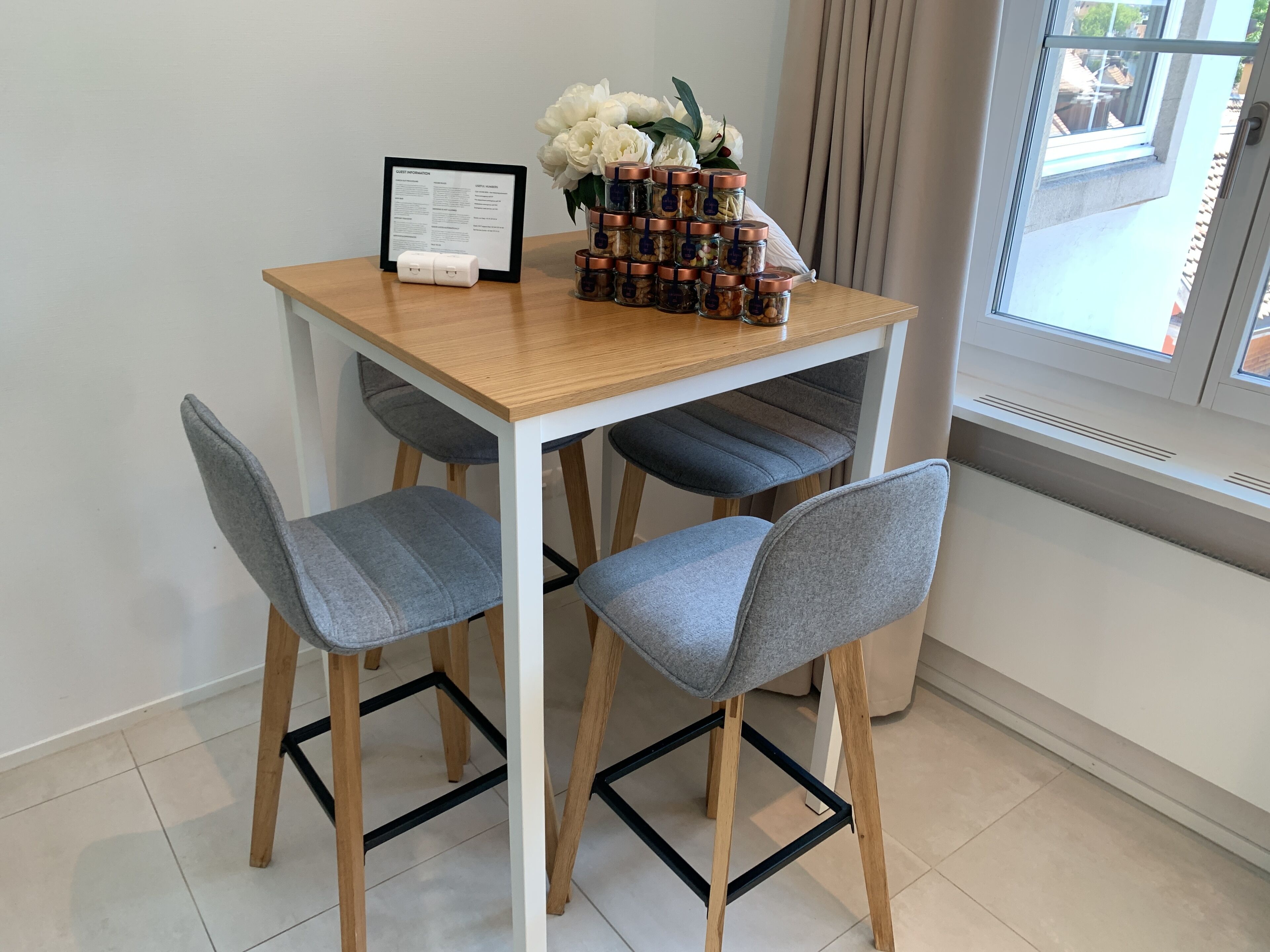 Een klein eetgedeelte met een houten tafel, grijs gestoffeerde stoelen, een lijst met het menu en een vaas met witte bloemen.