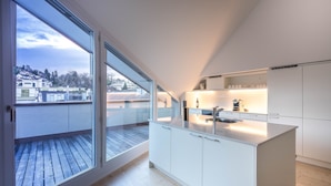 A modern kitchen with a large island, white cabinetry, and a view of the city through large windows. - Top city center Lion Monument (Lucerne)