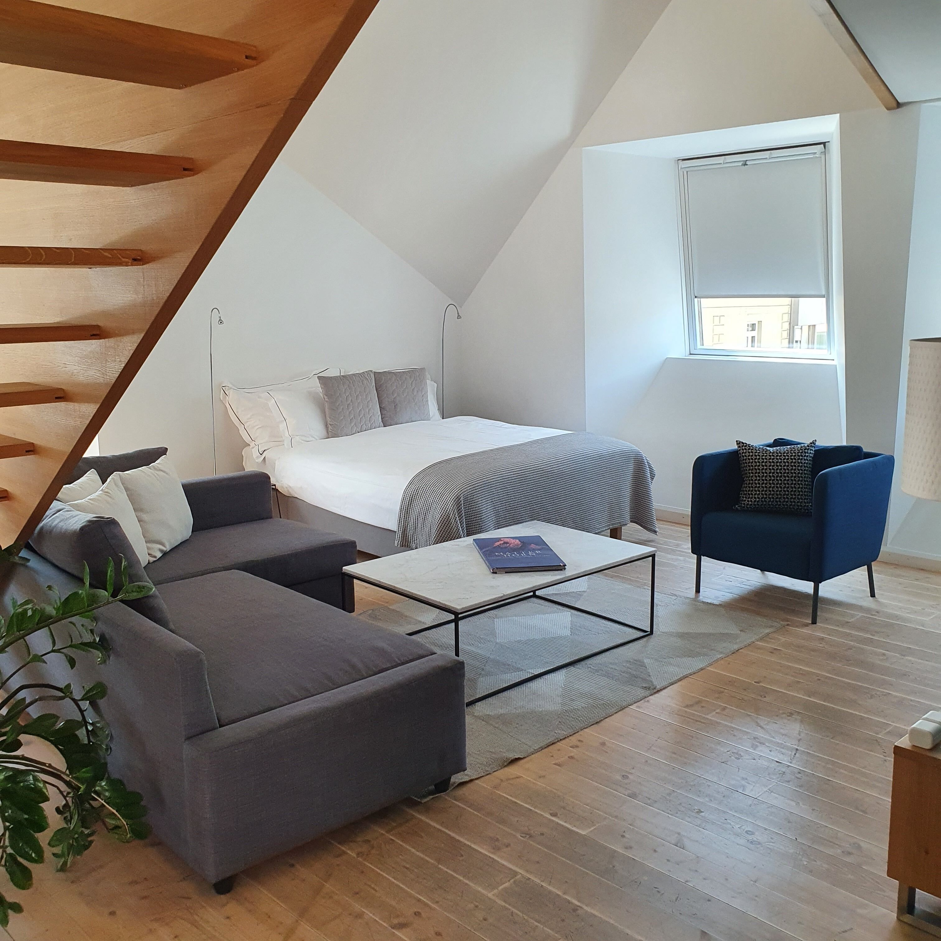 Camera da loft moderna con soffitto spiovente, un letto con piumone bianco, un divano grigio, un tavolino da caffè e una poltrona blu.