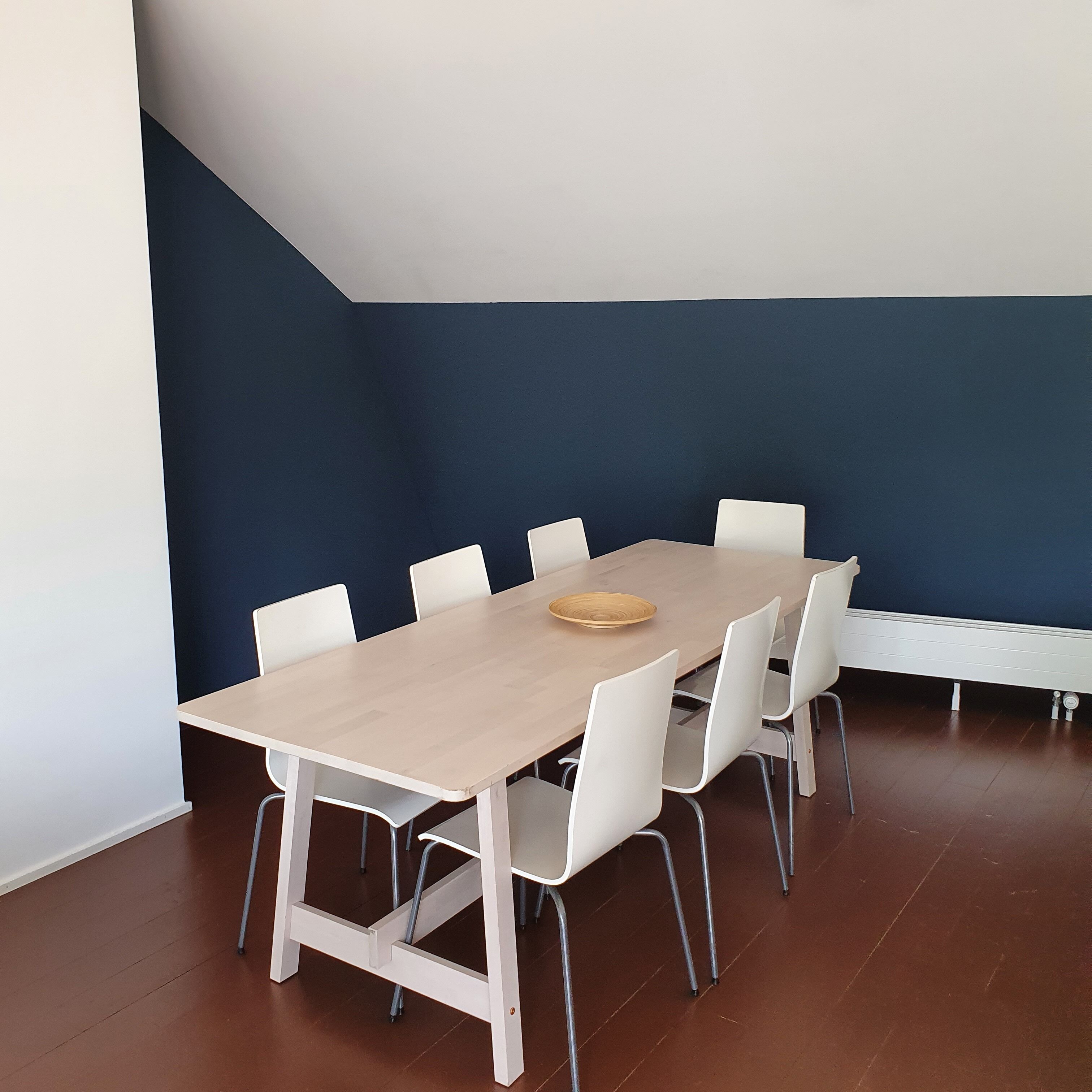 Une salle de conférence avec une longue table, des chaises blanches et un tableau en bois.