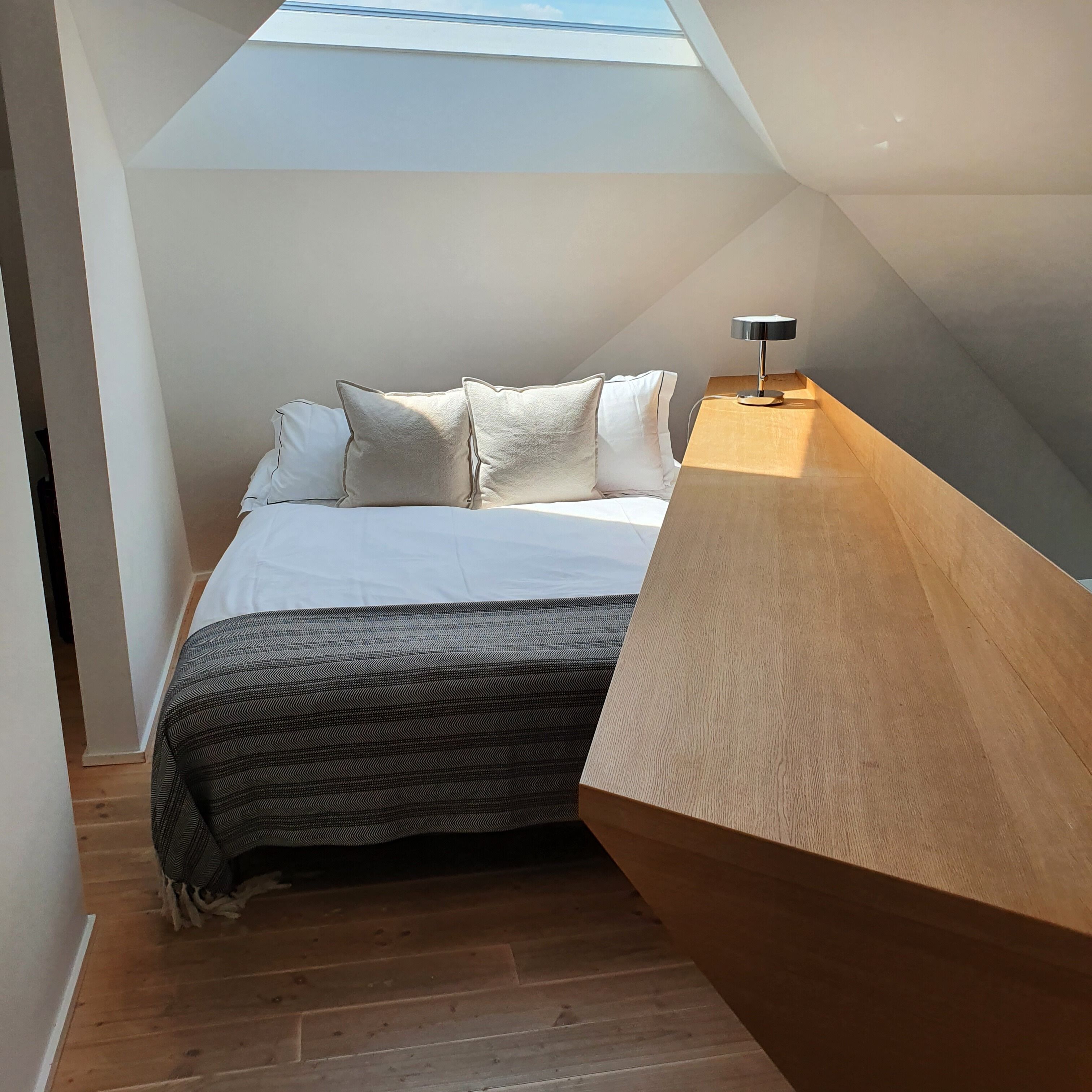 A bedroom with a skylight, a bed with white bedding, a patterned throw, and a wooden bedside table with a lamp.