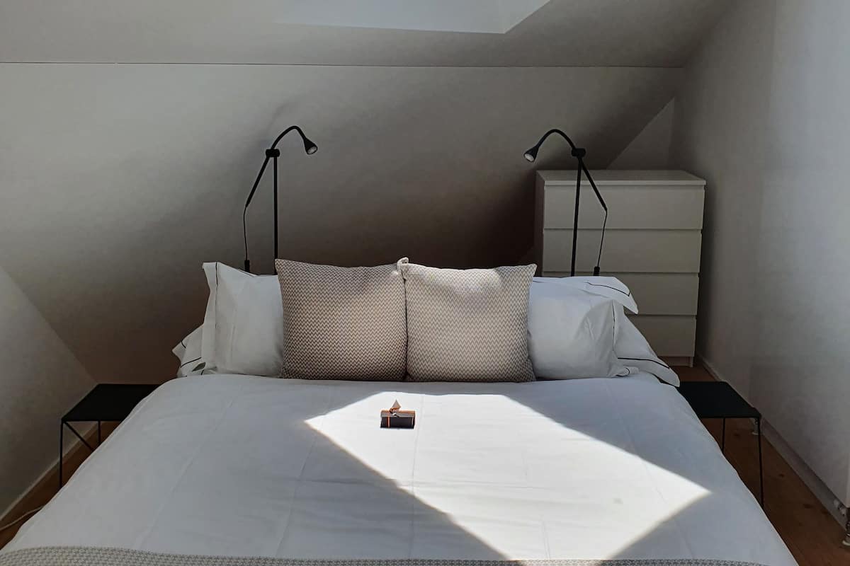 Un dormitorio con tragaluz, una cama con ropa blanca, dos mesitas de noche y un buró.