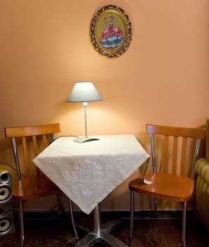 Ein kleiner Essbereich mit einem Tisch, der mit einer weißen Tischdecke bedeckt ist, zwei Holzstühlen, einer einzelnen Tischlampe und einem gerahmten Bild an der Wand.