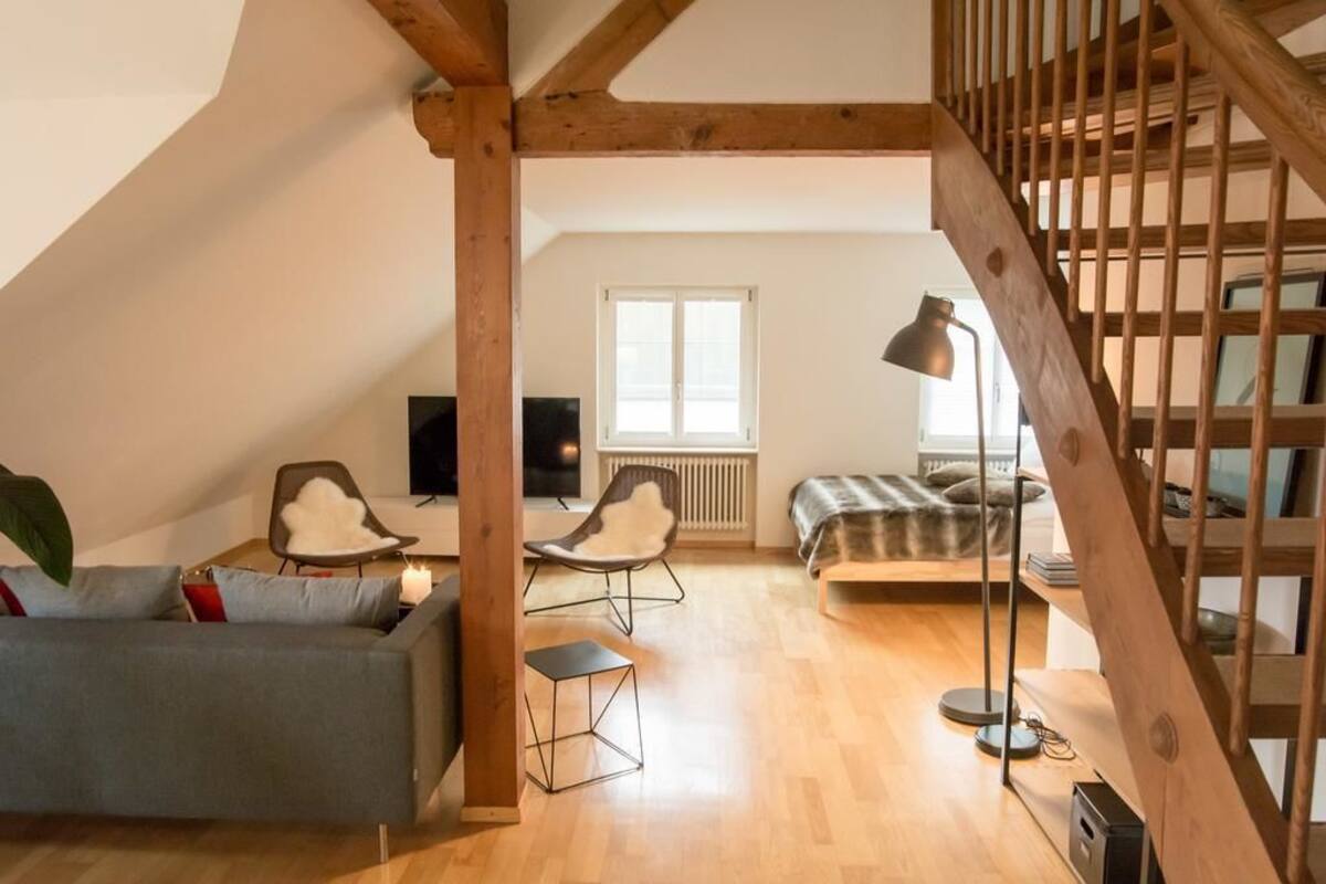 A loft-style room with a wooden staircase, a sofa, two chairs, a small table, and a bed.