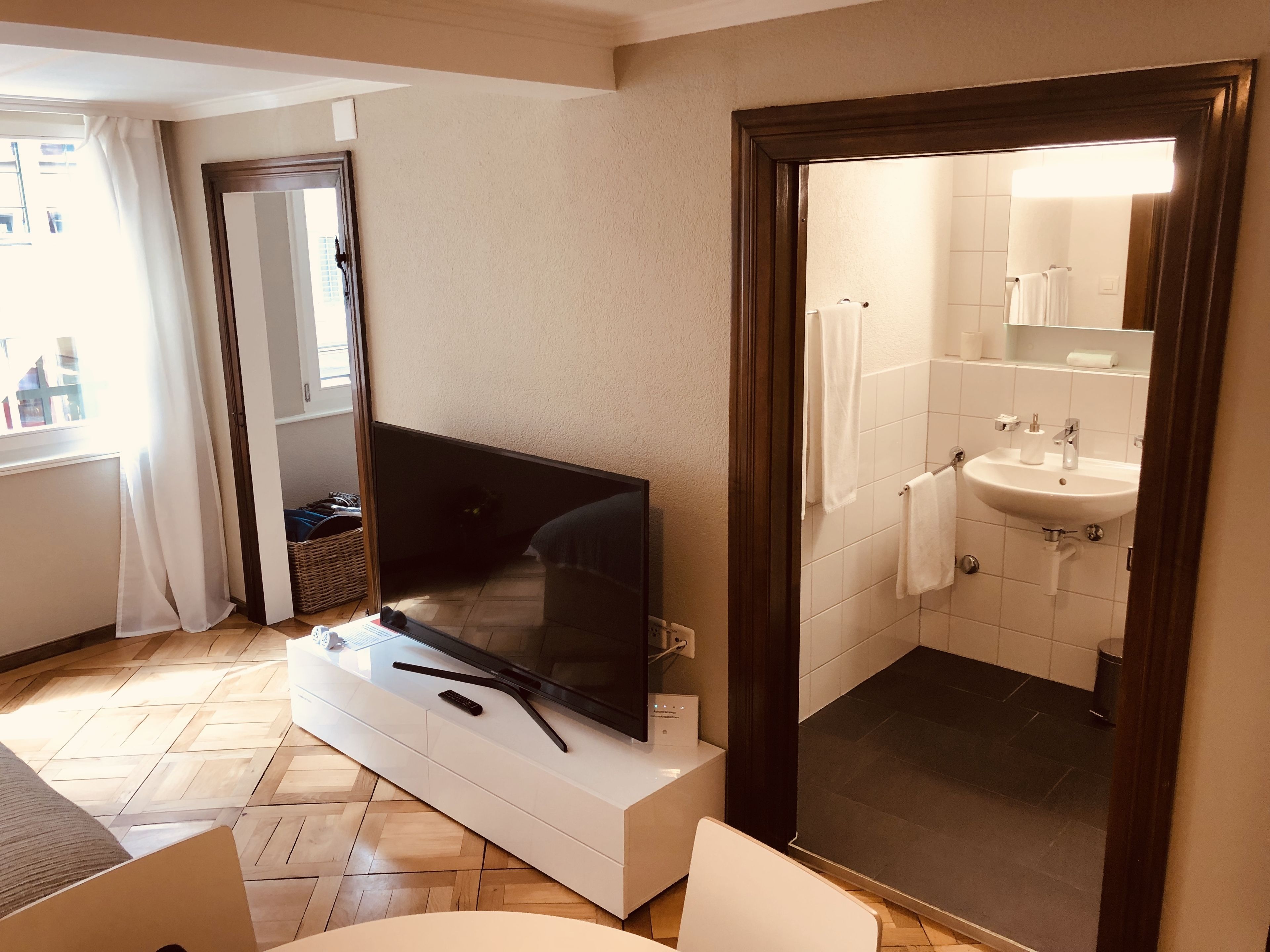 Habitación de hotel moderna con un televisor de pantalla plana sobre una consola blanca, piso de madera, un espejo grande y un baño con lavamanos y toallero.