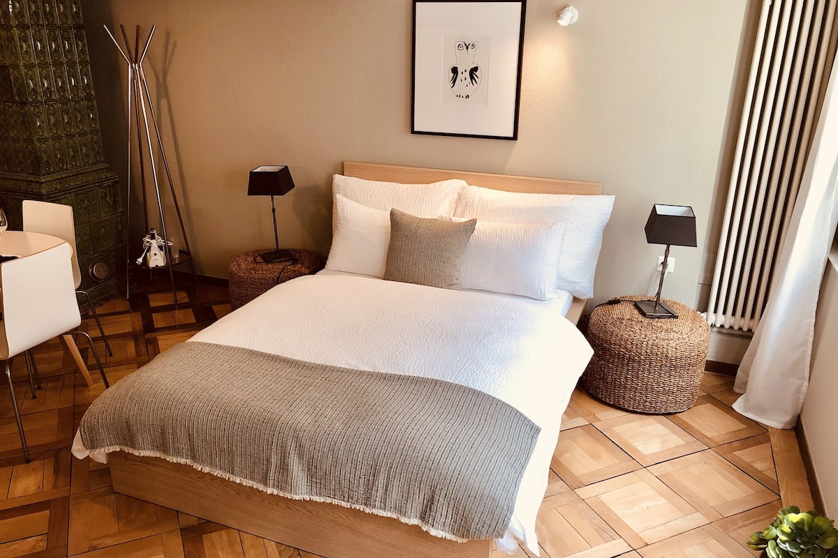 A neatly arranged bedroom with a large bed, a chair, a small table, a lamp, a framed picture, and a radiator.