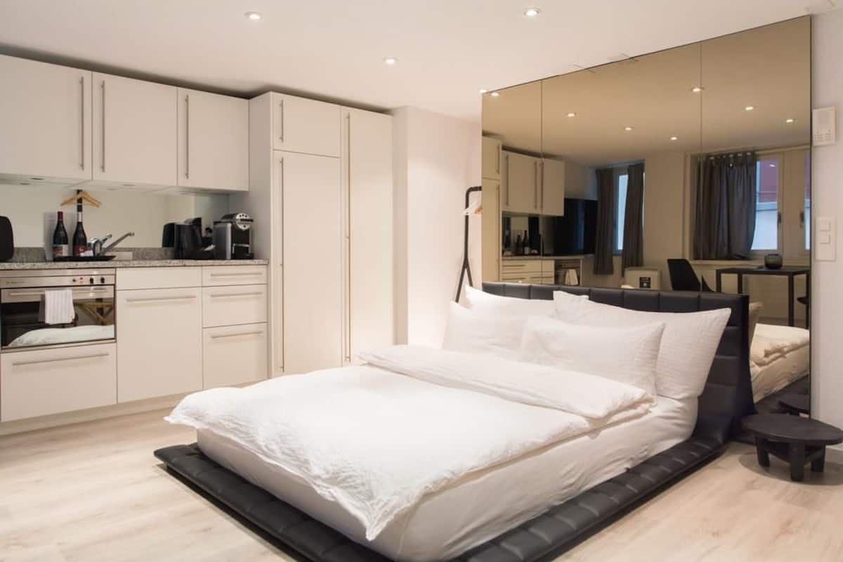 A modern bedroom with a large bed, a built-in wardrobe, a small kitchen area, and a large mirror reflecting the room.