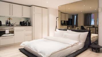 A modern bedroom with a large bed, a built-in wardrobe, a small kitchen area, and a large mirror reflecting the room.