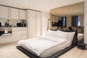 Un dormitorio moderno con una cama grande, un armario empotrado, una pequeña cocina y un espejo grande que refleja la habitación.