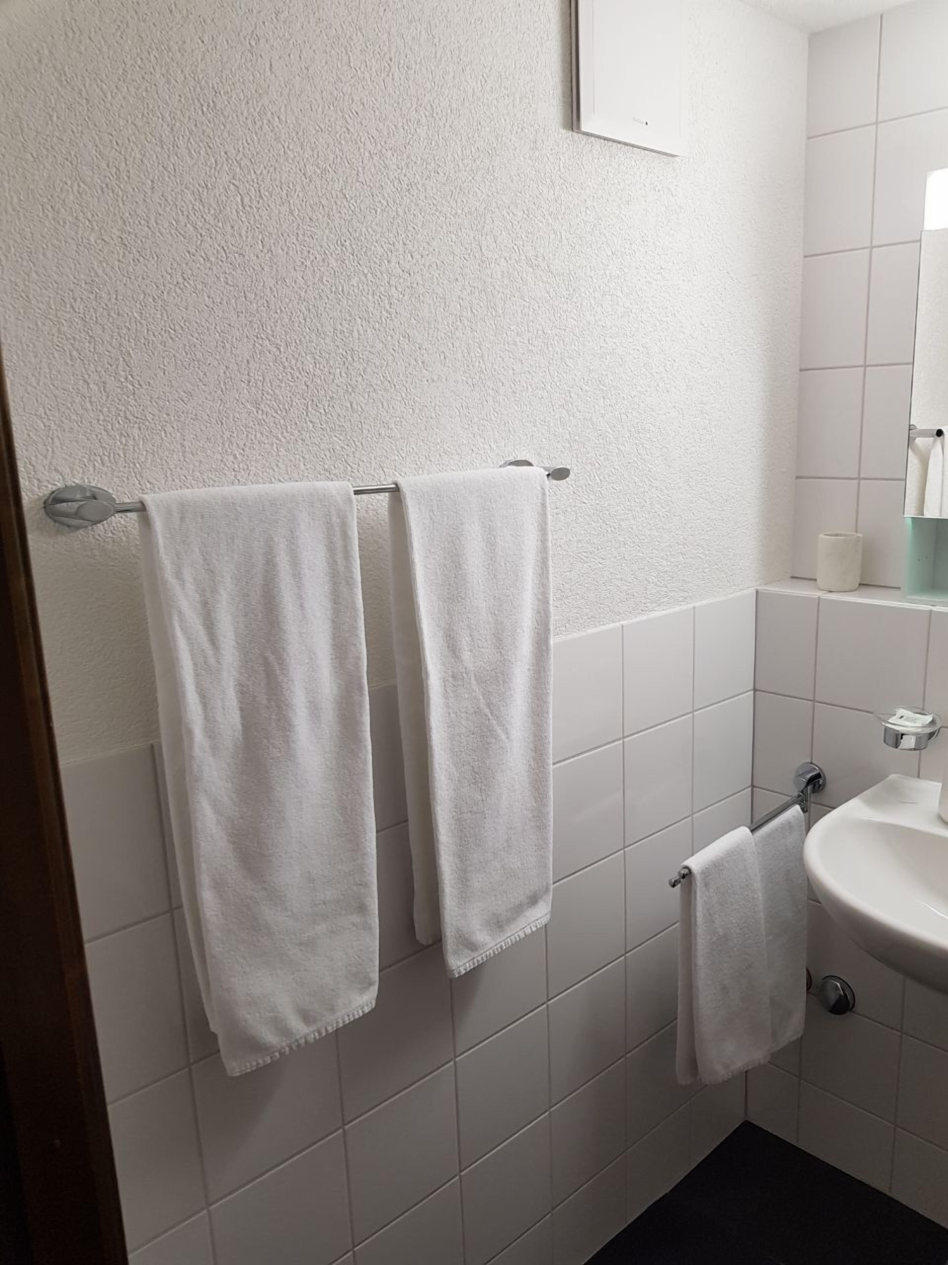 Une salle de bain avec des murs recouverts de carreaux blancs, un lavabo blanc et deux serviettes blanches suspendues à un porte-serviettes.