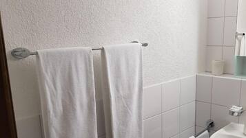 Une salle de bain avec des murs recouverts de carreaux blancs, un lavabo blanc et deux serviettes blanches suspendues à un porte-serviettes.