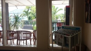 Una habitación con una mesa y sillas de madera, una ventana que ofrece vistas a palmeras y una cerca, y un cartel que dice "SILAHKAN DIBACA".
