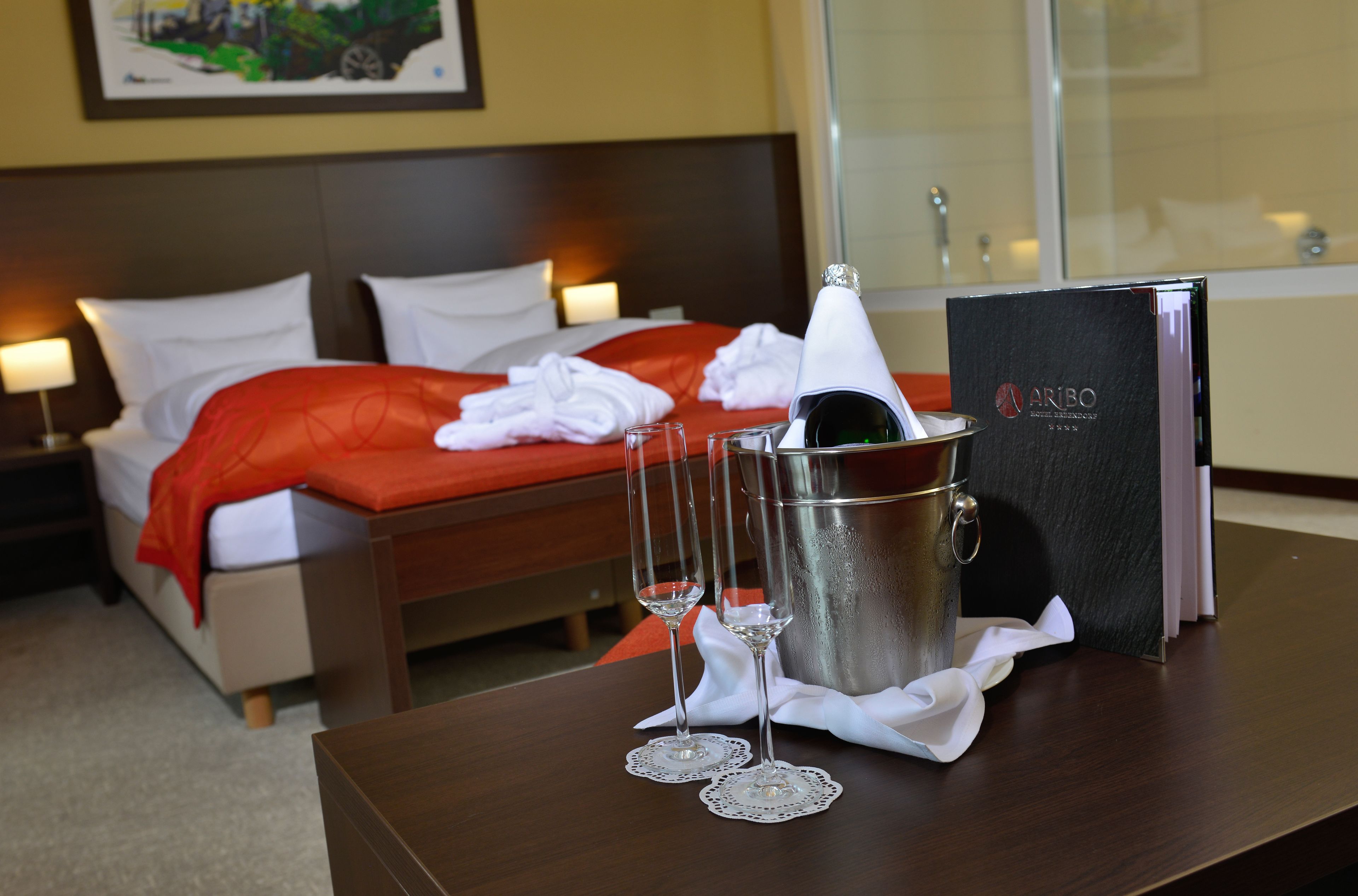 A hotel room with a neatly made bed, a minibar, and two glasses of champagne on the table.