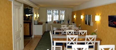 Un comedor con mesas de madera y sillas blancas, una cocina al fondo y vistas al exterior a través de amplios ventanales.