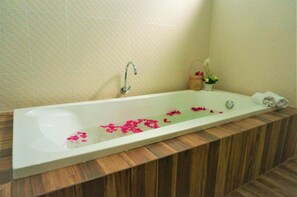 A rectangular bathtub with a wooden base, a white wall with a textured pattern, and a small basket with flowers. - Ruenthai Boutique (Sa Khu)