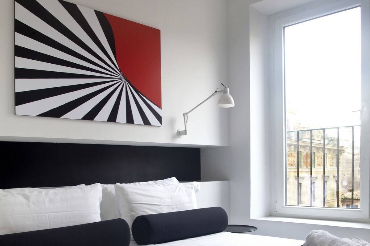 A modern hotel room with a large bed, white and black bedding, a red and black abstract painting, a white desk lamp, and a view of the city through a large window.
