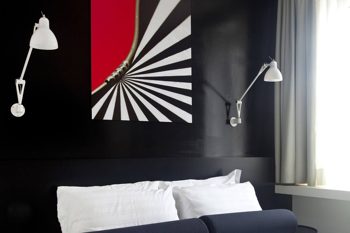 A modern hotel room with a large bed, two bedside lamps, a laptop, and a newspaper on the bed.