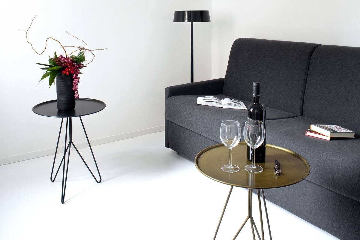 A modern living room with a grey sofa, a round coffee table with two wine glasses and a bottle, a black floor lamp, a white pendant light, and a wall art piece.