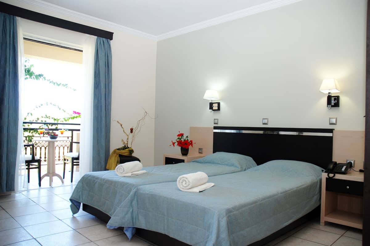 A hotel room with two beds, a small table with chairs, a balcony with plants, and two wall-mounted lamps.