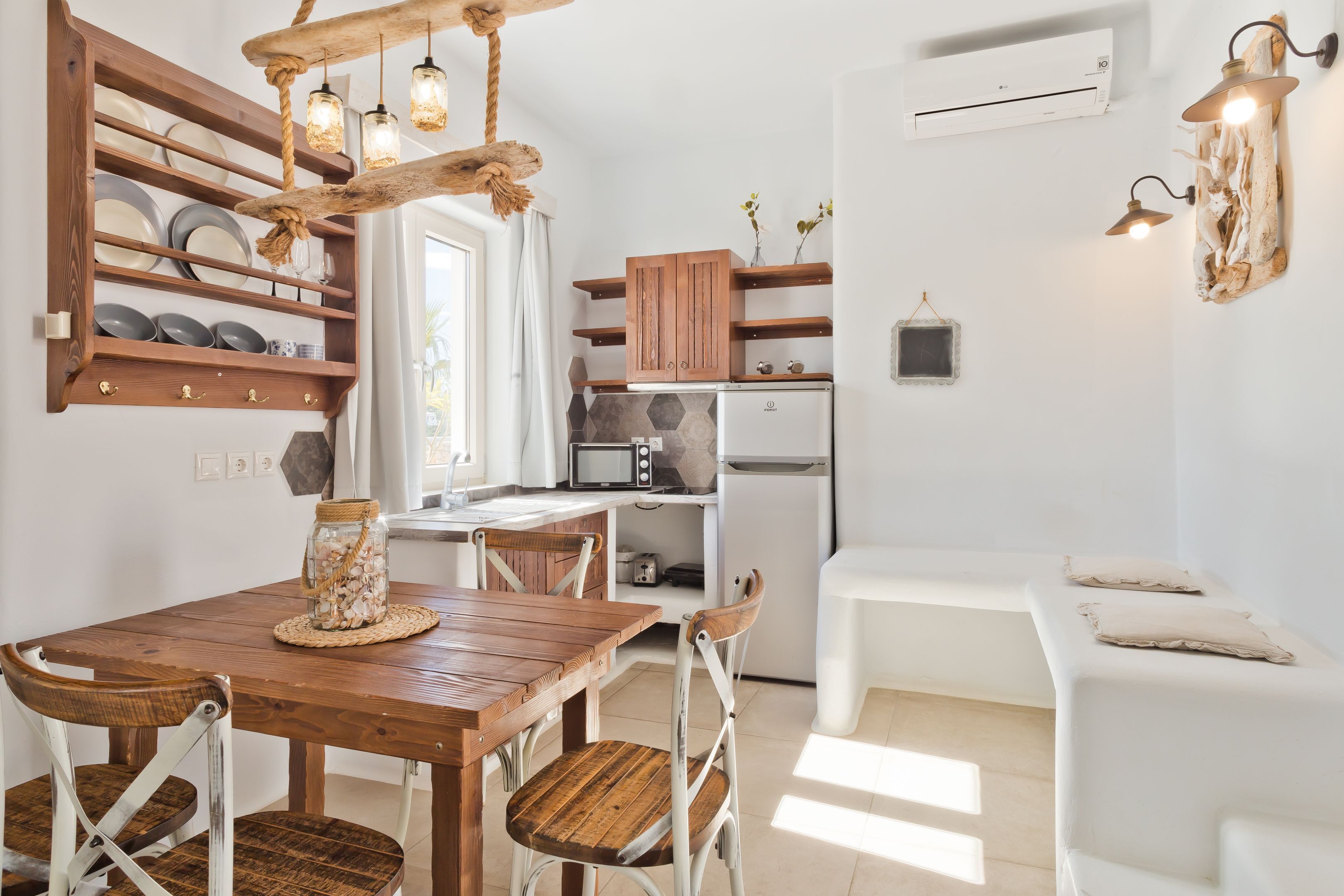 Una cocina compacta con armarios de madera, una mesa de comedor y sillas. Hay un banco blanco en la esquina y una lámpara colgante sobre la mesa.
