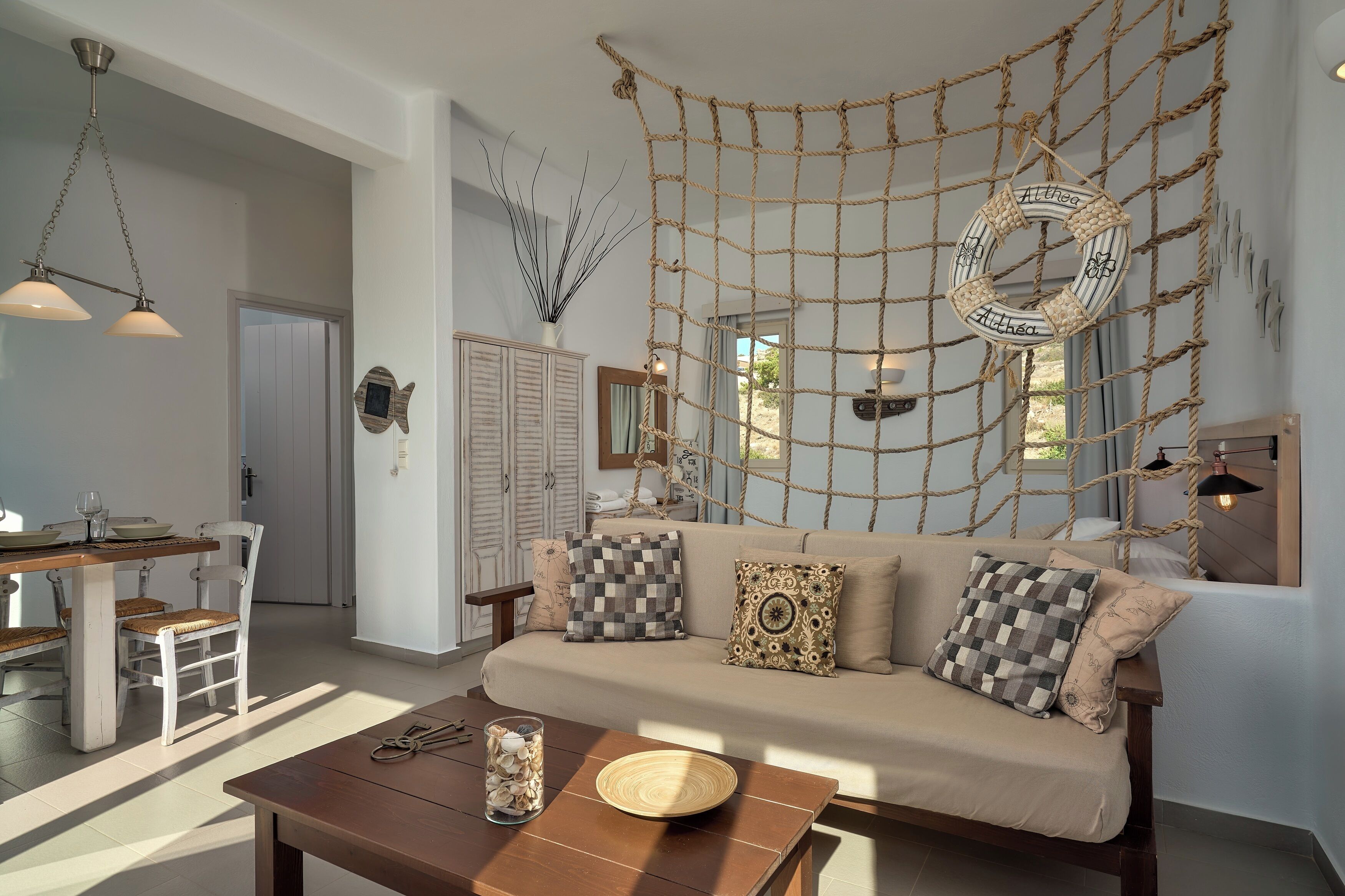 A cozy living room with a sofa, coffee table, and a rope wall hanging. There is a dining area with a table and chairs, and a kitchen in the background.