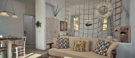 A cozy living room with a sofa, coffee table, and a rope wall hanging. There is a dining area with a table and chairs, and a kitchen in the background.