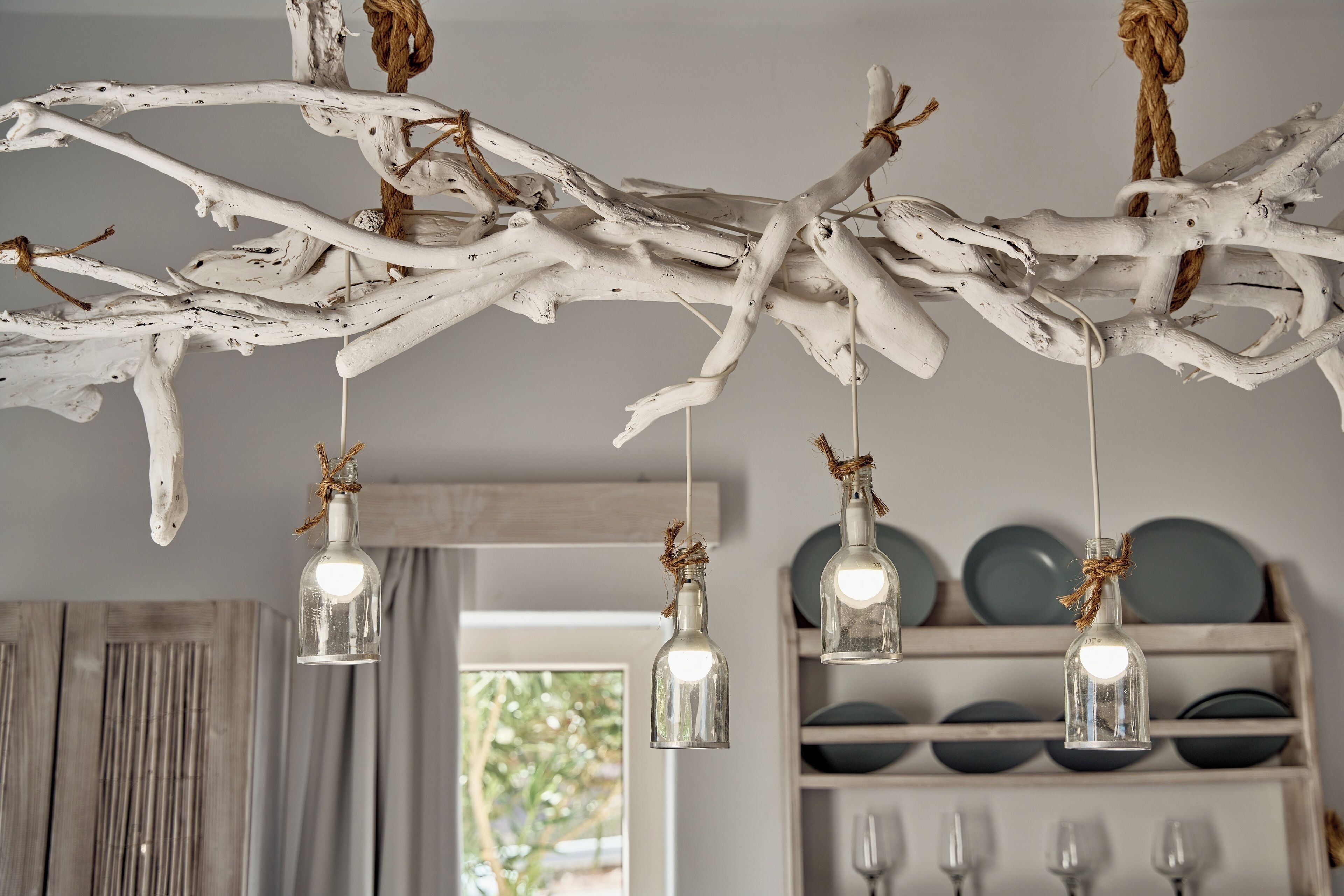 Un rustico lampadario di rami bianchi con lampadine in vetro, appeso in una stanza con uno scaffale di legno sullo sfondo.