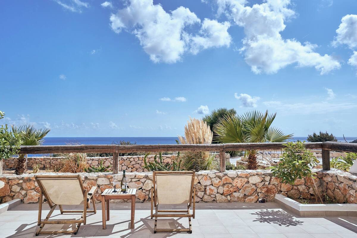 A terrace with two wooden chairs and a table, overlooking a stone wall and the sea.