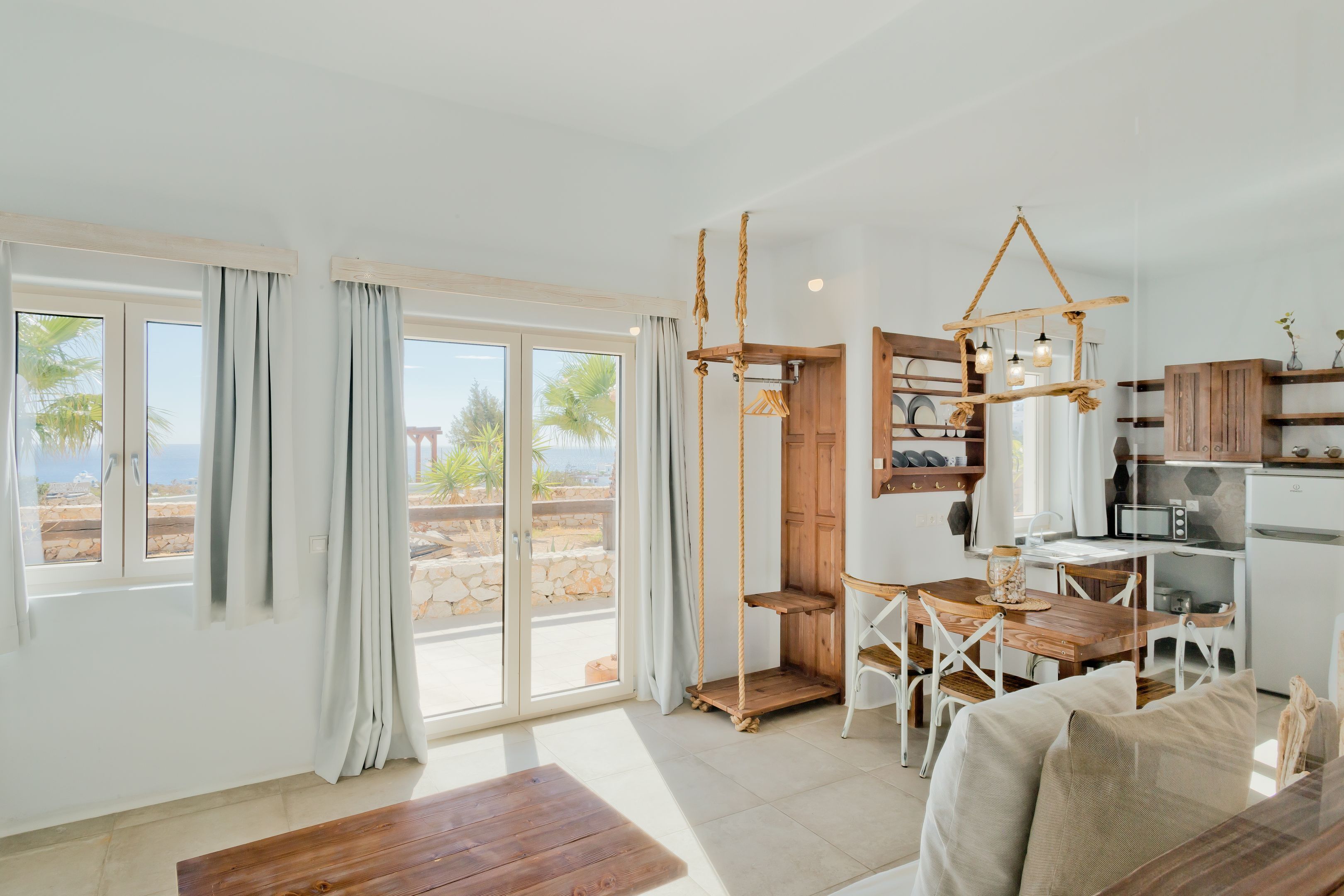 A spacious living area with a wooden dining table, white chairs, and a sofa. There is a kitchen area with wooden cabinets and a microwave. Large windows provide a view of the outdoors.