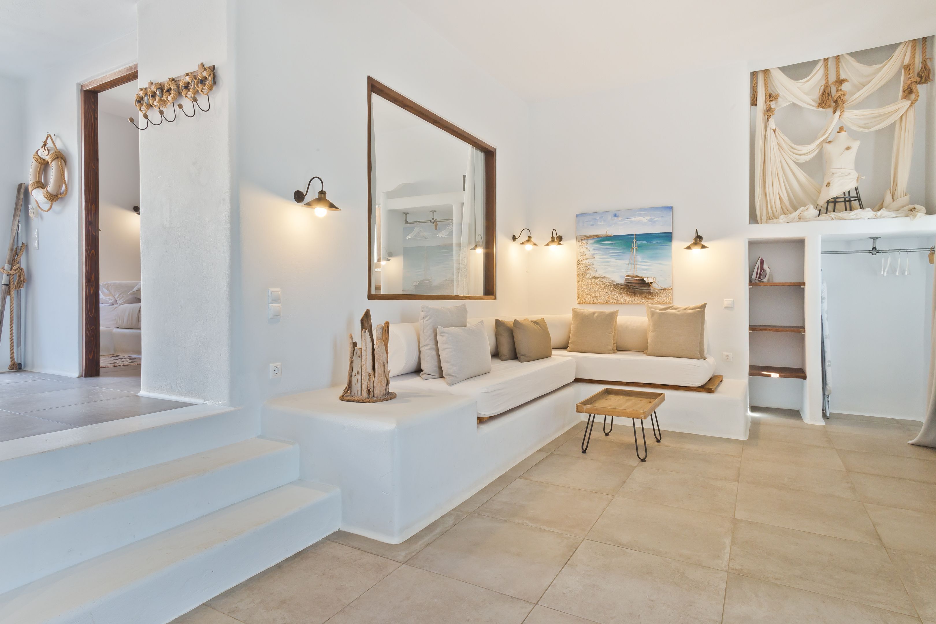 A modern living room with a white sofa, a coffee table, and a painting of a beach scene. There is a staircase leading to another room, and a wooden door with a coat rack.