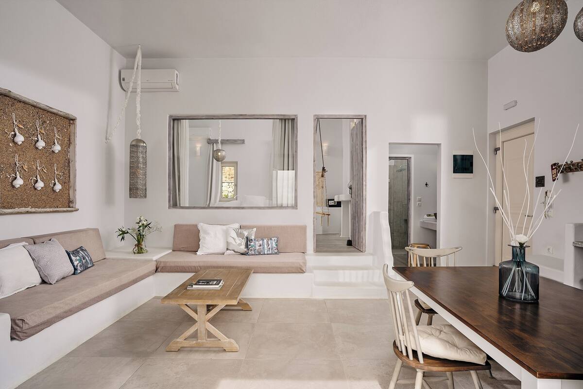 A modern living room with a sofa, coffee table, and dining table. There is a large window, a mirror, and a hanging light fixture.
