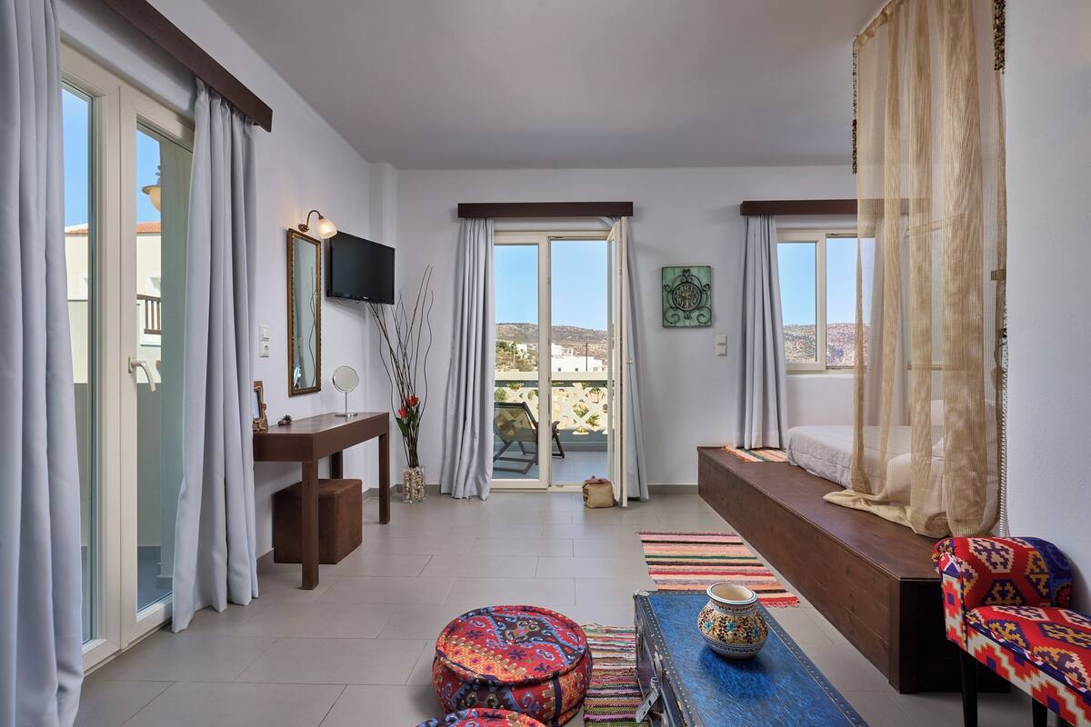 A spacious living area with a sofa, ottoman, and a coffee table. There is a TV mounted on the wall, a window with curtains, and a view of the outdoors.