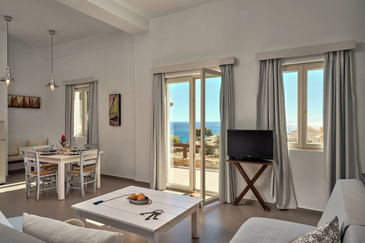 A modern living room with a dining area, a TV, and large windows with ocean views.