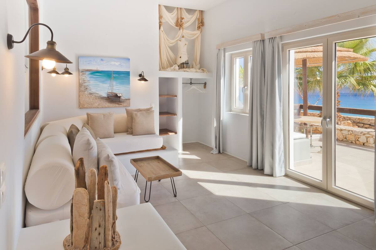 A modern living room with a white sofa, a wooden coffee table, and a painting of a beach scene. There is a sliding glass door leading to an outdoor area with a view of the ocean and palm trees.