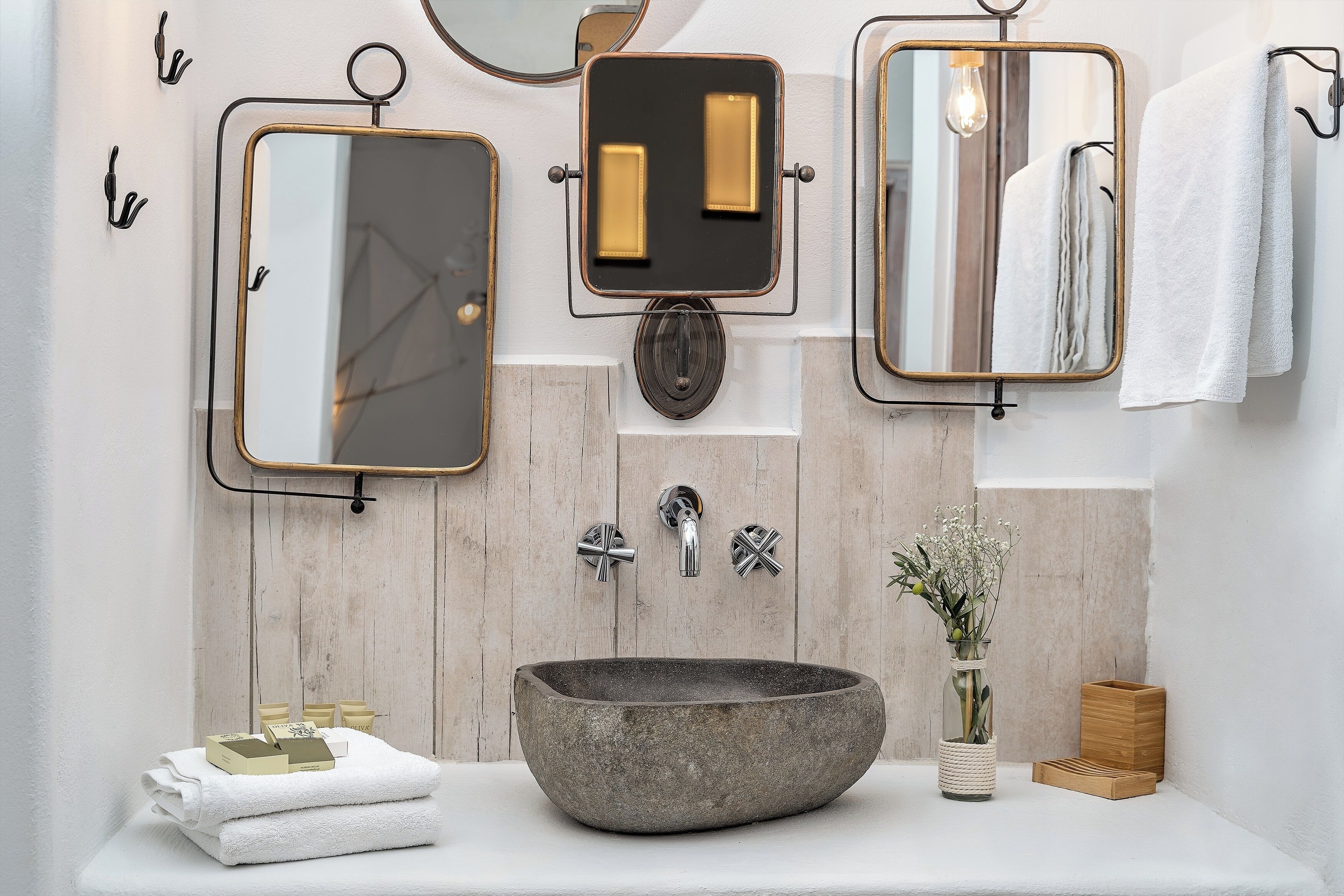 A bathroom with a stone sink, two mirrors, a towel rack, and a vase with flowers.