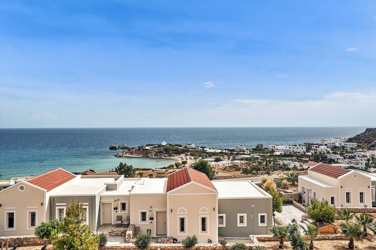 A coastal residential area with white buildings, a red-roofed house in the foreground, and a view of the sea and rocky coastline in the background.