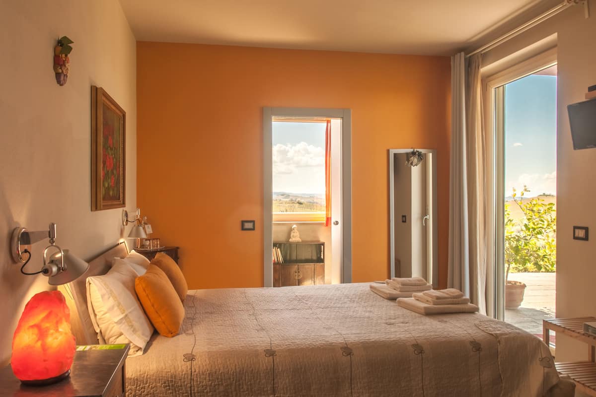 A bedroom with a bed, a nightstand, a heater, a door leading to a balcony, and a view of the outdoors.