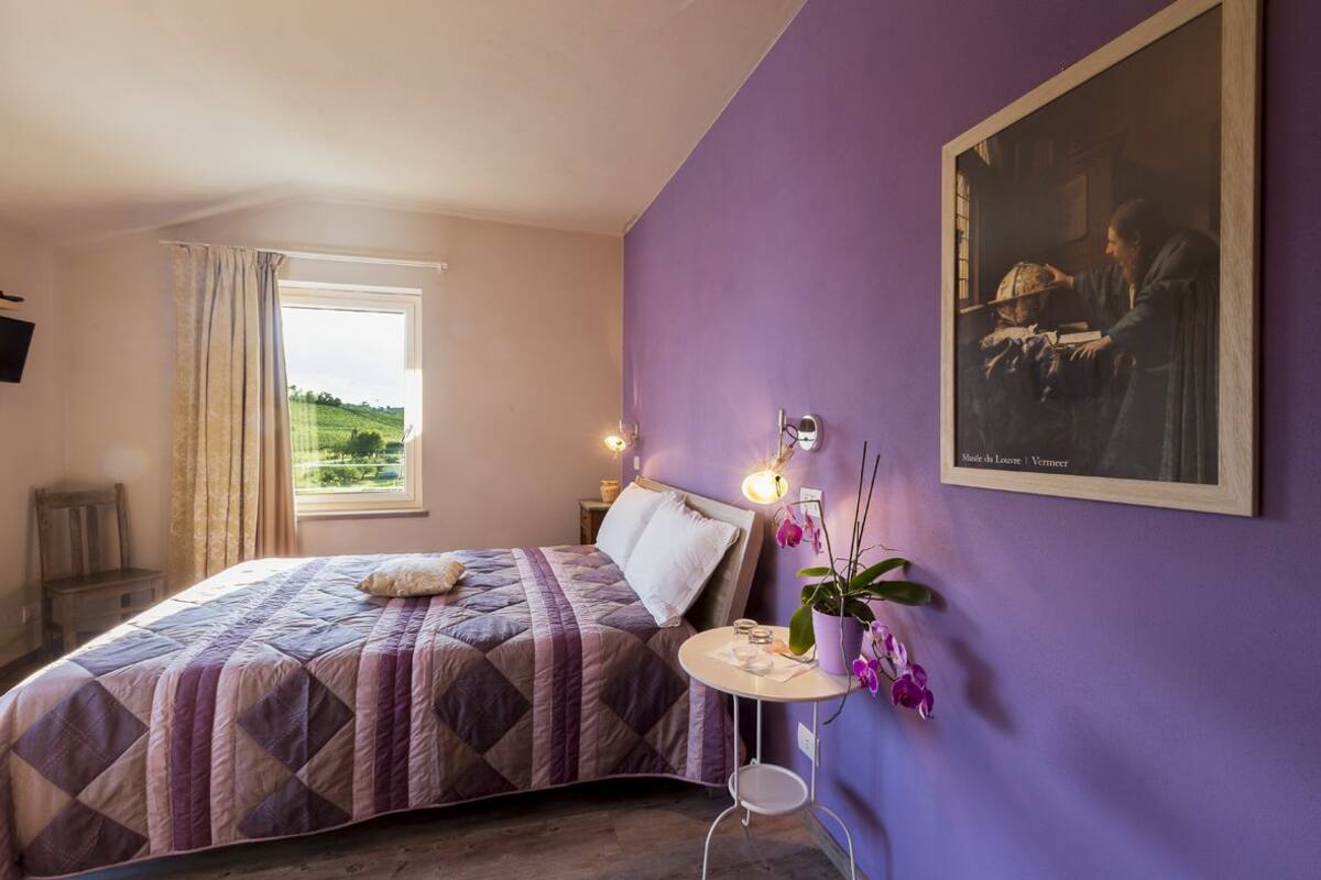 A bedroom with a bed, a window with curtains, a small table with a plant, and a painting on the wall.