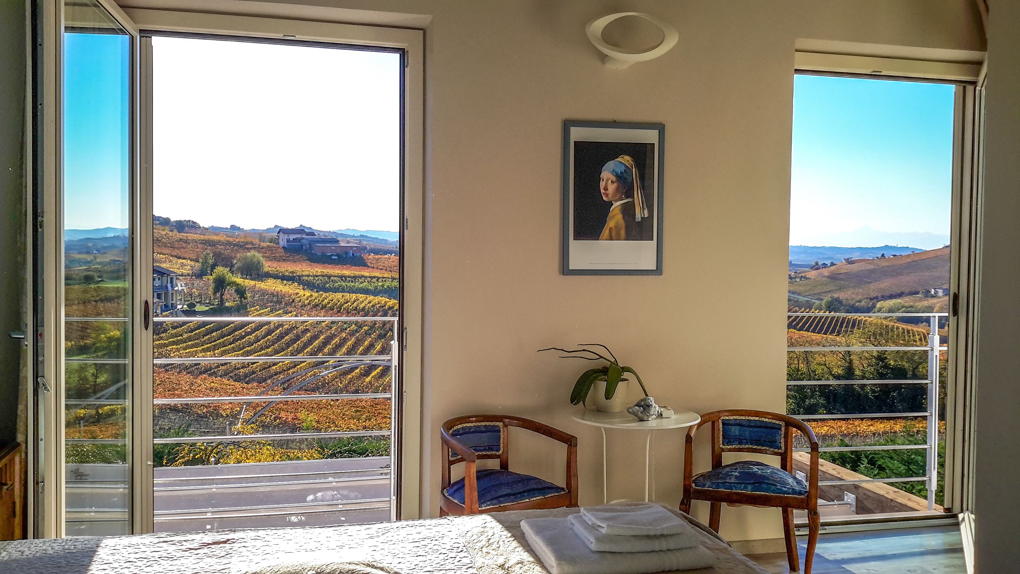 A balcony with a view of vineyards, a framed picture on the wall, and two chairs.
