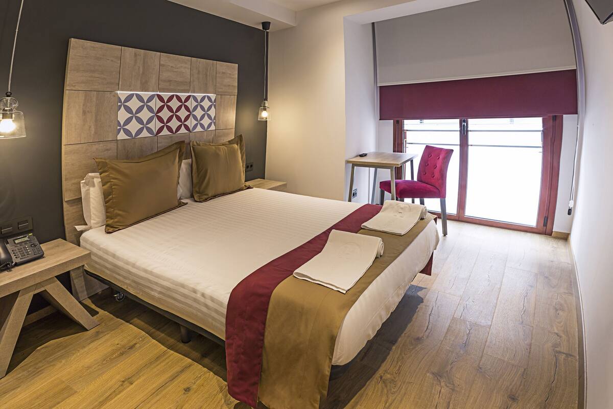 A hotel room with a large bed, a desk with a telephone, a chair, and a window with a view.