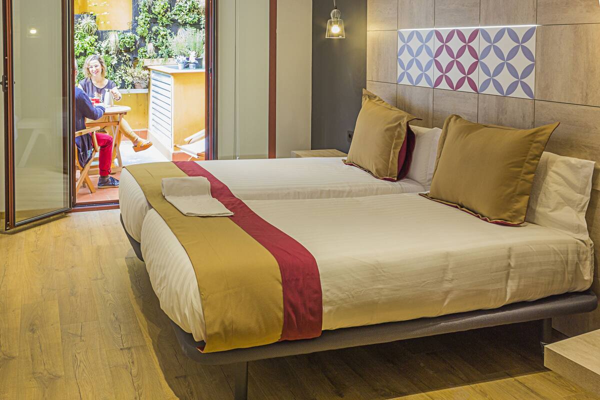 A double bed with a beige and red bedspread, a wooden headboard, and a wall-mounted light fixture.