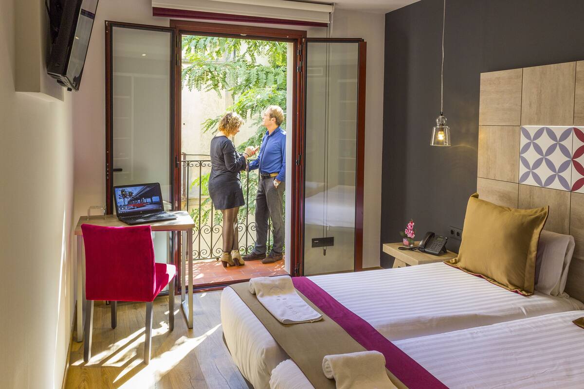 A hotel room with a bed, a desk with a laptop, a red chair, and a view of the outside.